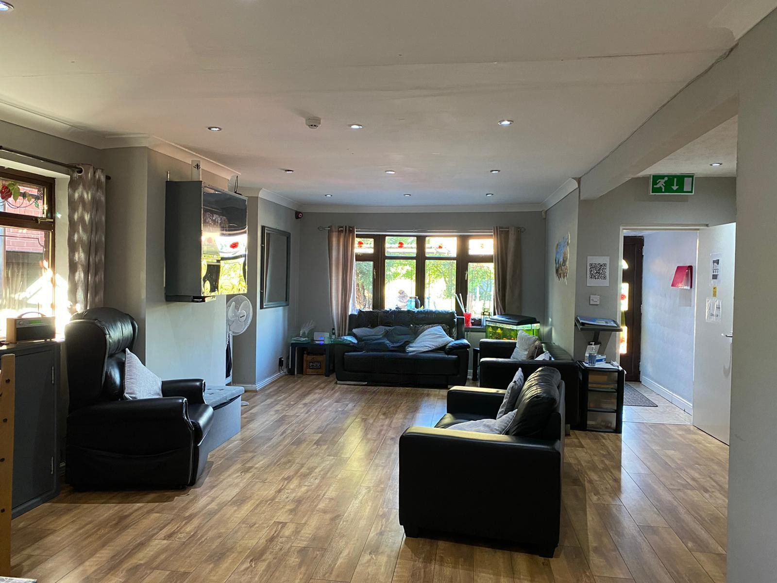A spacious lounge at Harden Residential Home with wooden flooring, black armchairs, a sofa, a television on the wall, and large windows letting in sunlight. A doorway leads to another well-lit room.