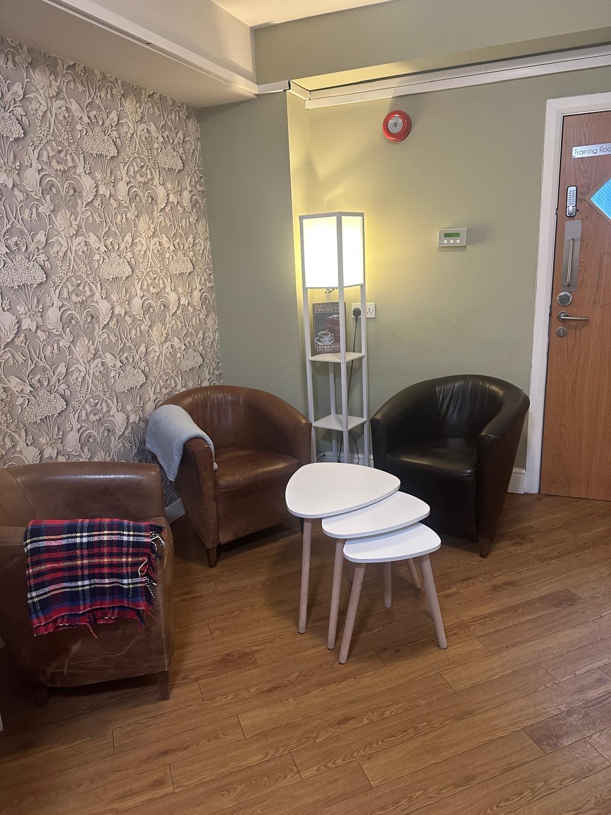 A cosy seating area at St. Anthony’s Care Home features two brown armchairs, a small stack of white tables, a lamp with shelves holding books, and a door. One chair has a checked blanket; the room has wooden flooring and patterned wallpaper.