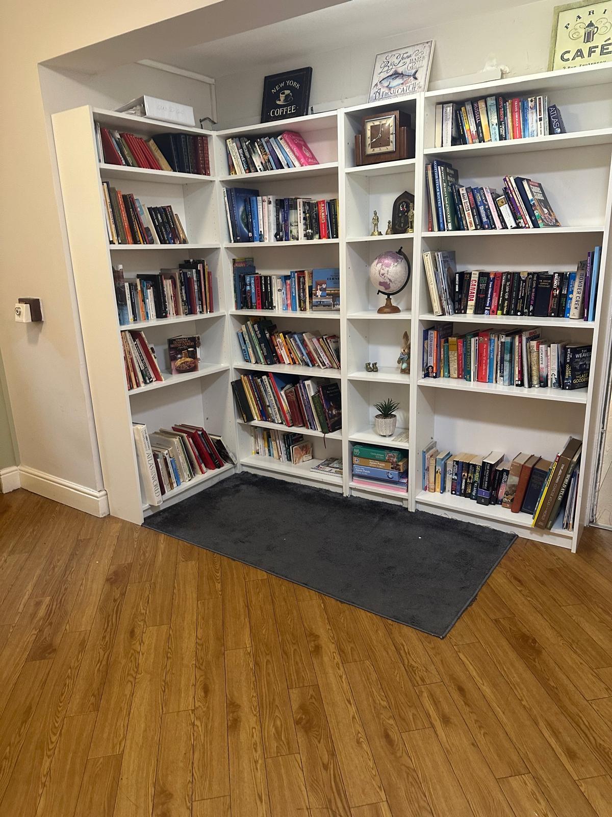 A cosy reading nook at St. Anthony’s Care Home features white corner bookshelves filled with books, a globe, and a small plant. A grey rug on the wooden floor adds comfort whilst signs and art hang above the shelves, creating a welcoming senior care space.