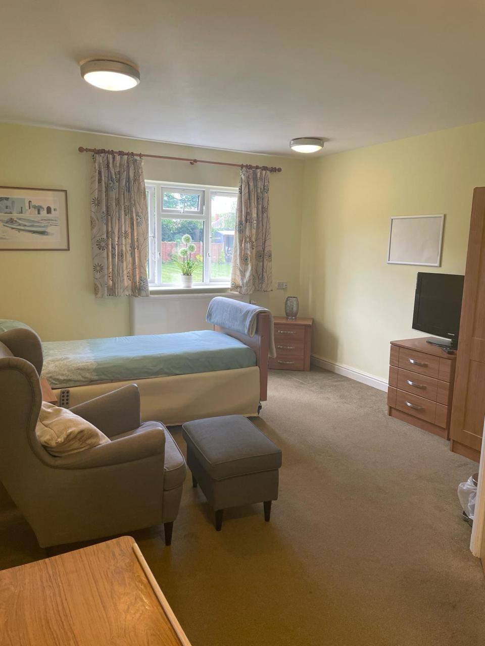 A tidy bedroom at St. Anthony’s Care Home features a single bed, armchair, ottoman, TV on a wooden chest of drawers, and a window with floral curtains—offering comfort and tranquillity in a welcoming care home environment.