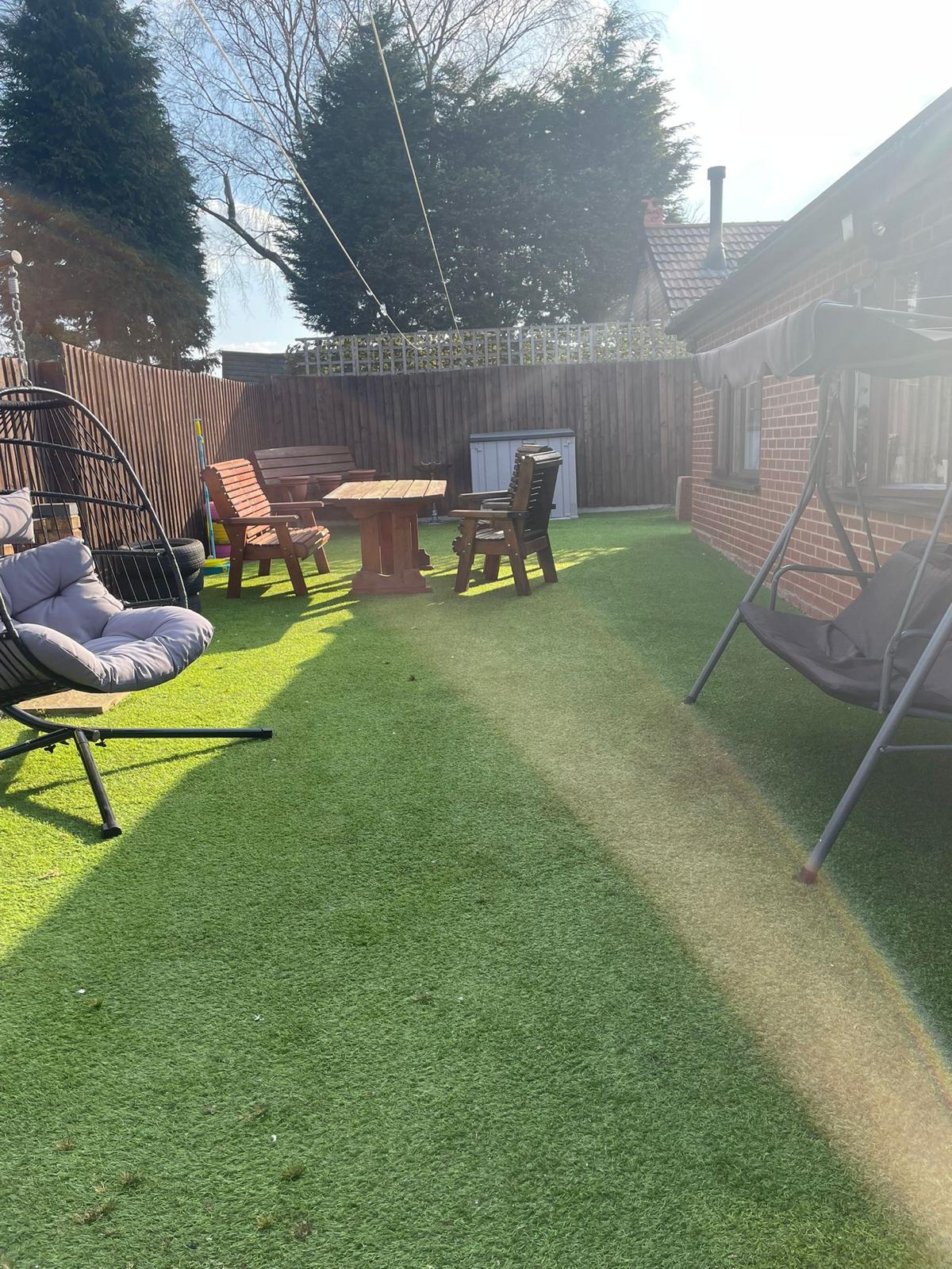 A sunny back garden at a Harden Residential Home features artificial grass, wooden outdoor furniture, a hanging chair, and a swing seat. Sunlight brightens the grass as trees and a wooden fence line the peaceful outdoor space.