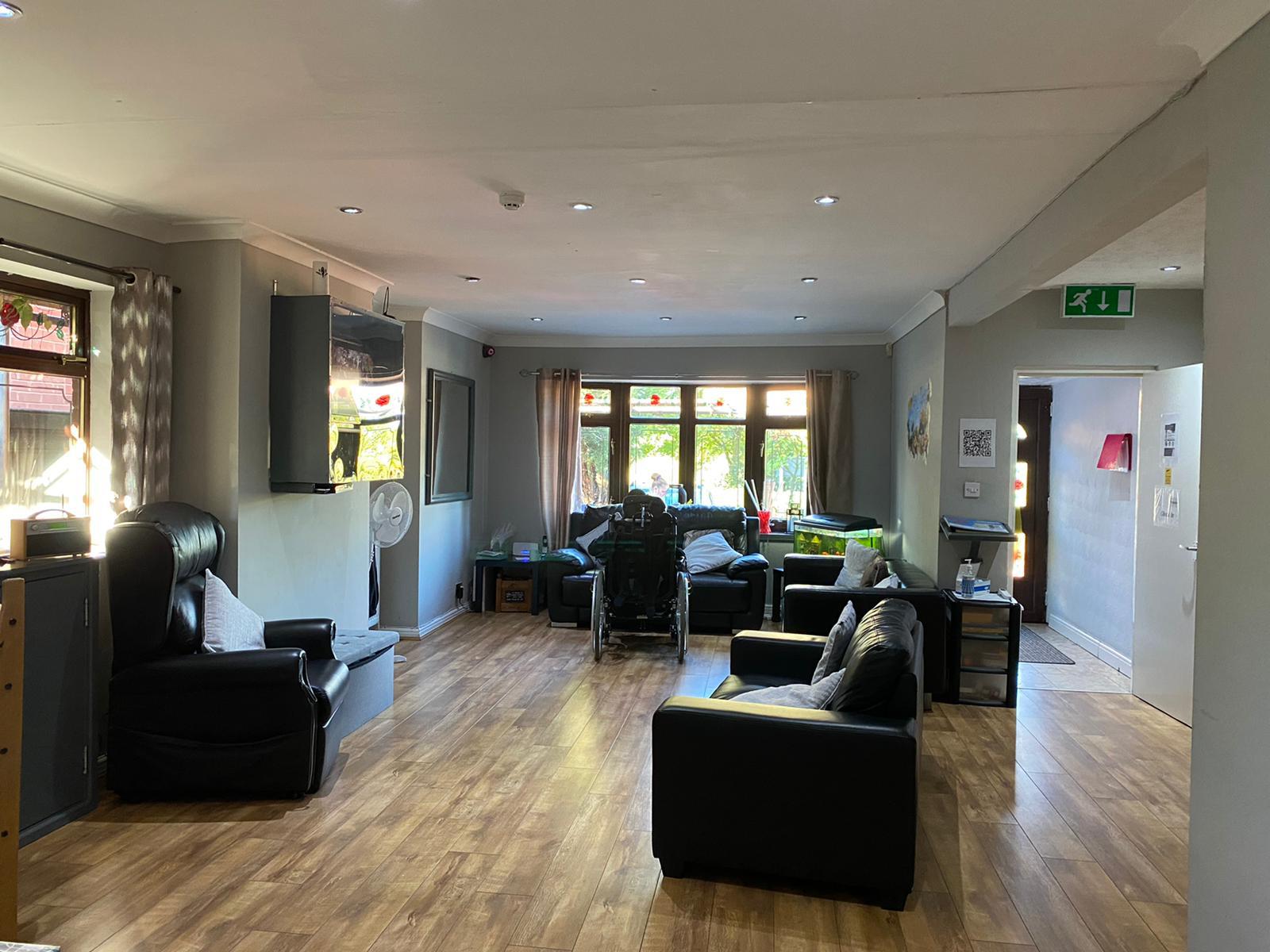 A spacious living room in Harden Residential Home features wooden flooring, black armchairs, and large windows filling the space with light. Plants, a wall-mounted TV, and a green emergency exit sign above a doorway complete this welcoming residential home.