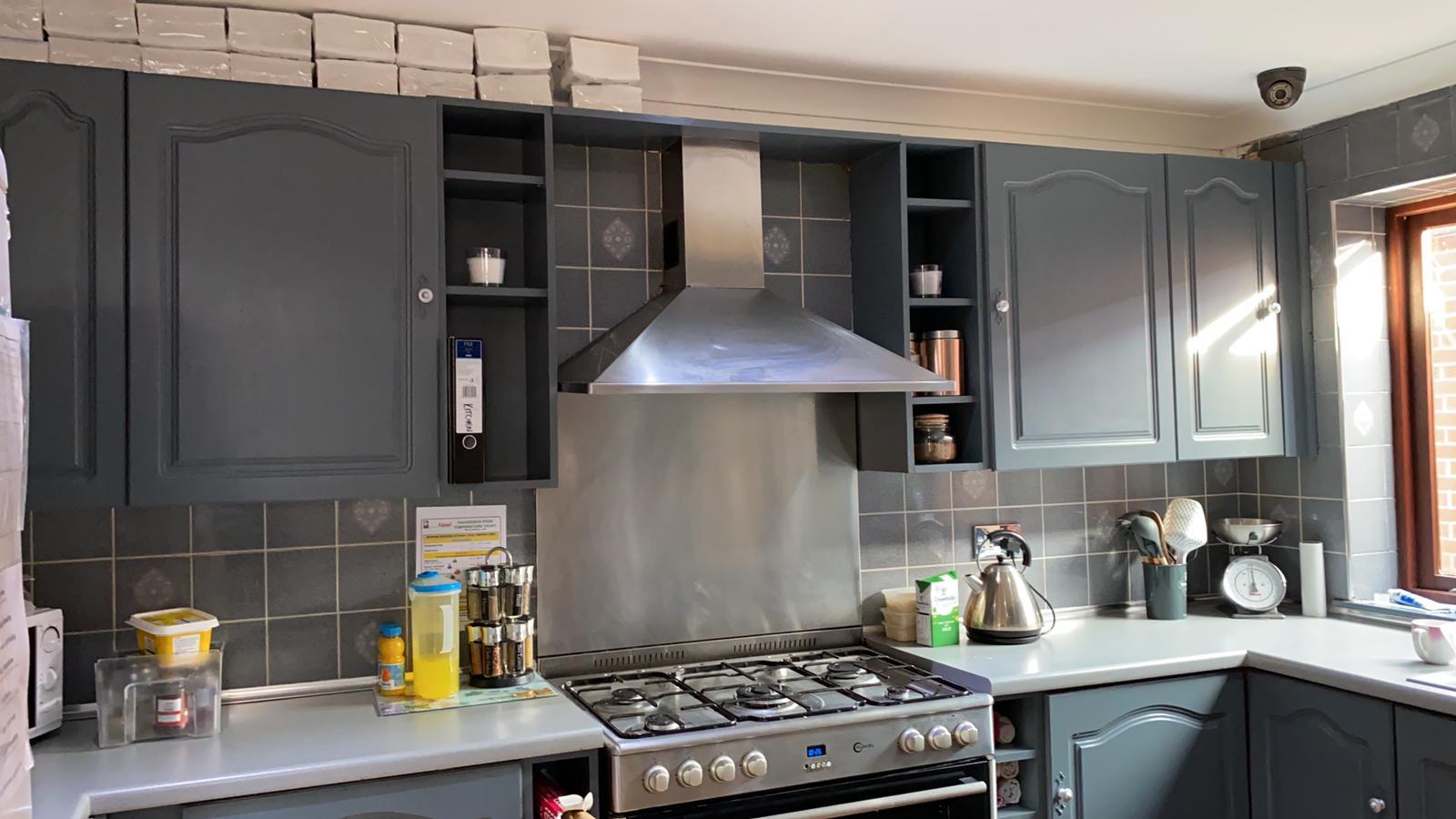 Modern kitchen in a residential home with dark grey cupboards, a stainless steel cooker and extractor hood, and a tiled splashback. Kitchen appliances, spices, and utensils are neatly arranged as sunlight hardens through the window on the right.