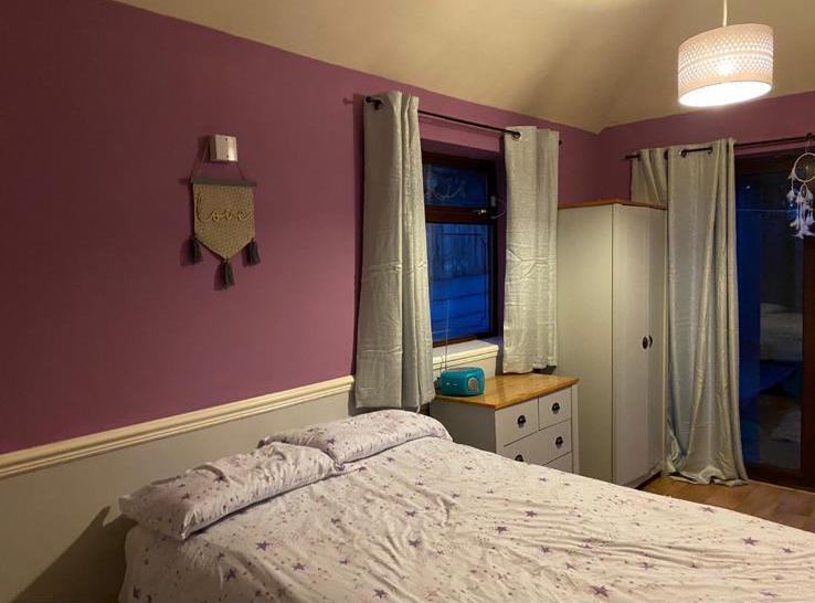 A cosy bedroom in a residential home with mauve and light grey walls, a bed with star-patterned bedding, a white wardrobe, wooden chest of drawers with blue radio, light curtains, and a ceiling lamp creates an inviting space to relax and strengthen your dreams.