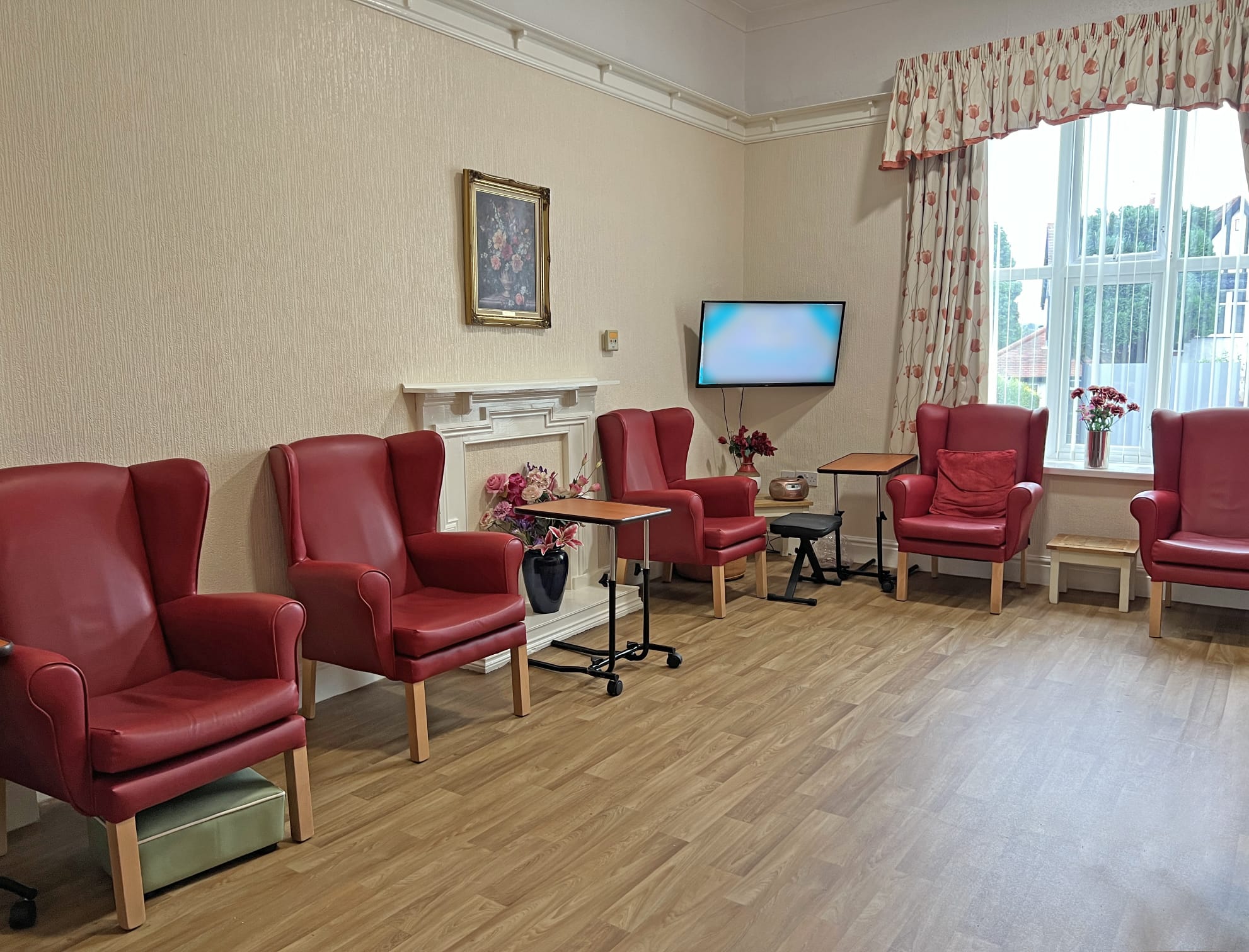 A cosy room at Park Lane Care Home features red armchairs in a row along the wall, small tables with flowers, a painting above the mantelpiece, flat-screen TV, large window with patterned curtains, and wood-style flooring—perfect for elderly care.