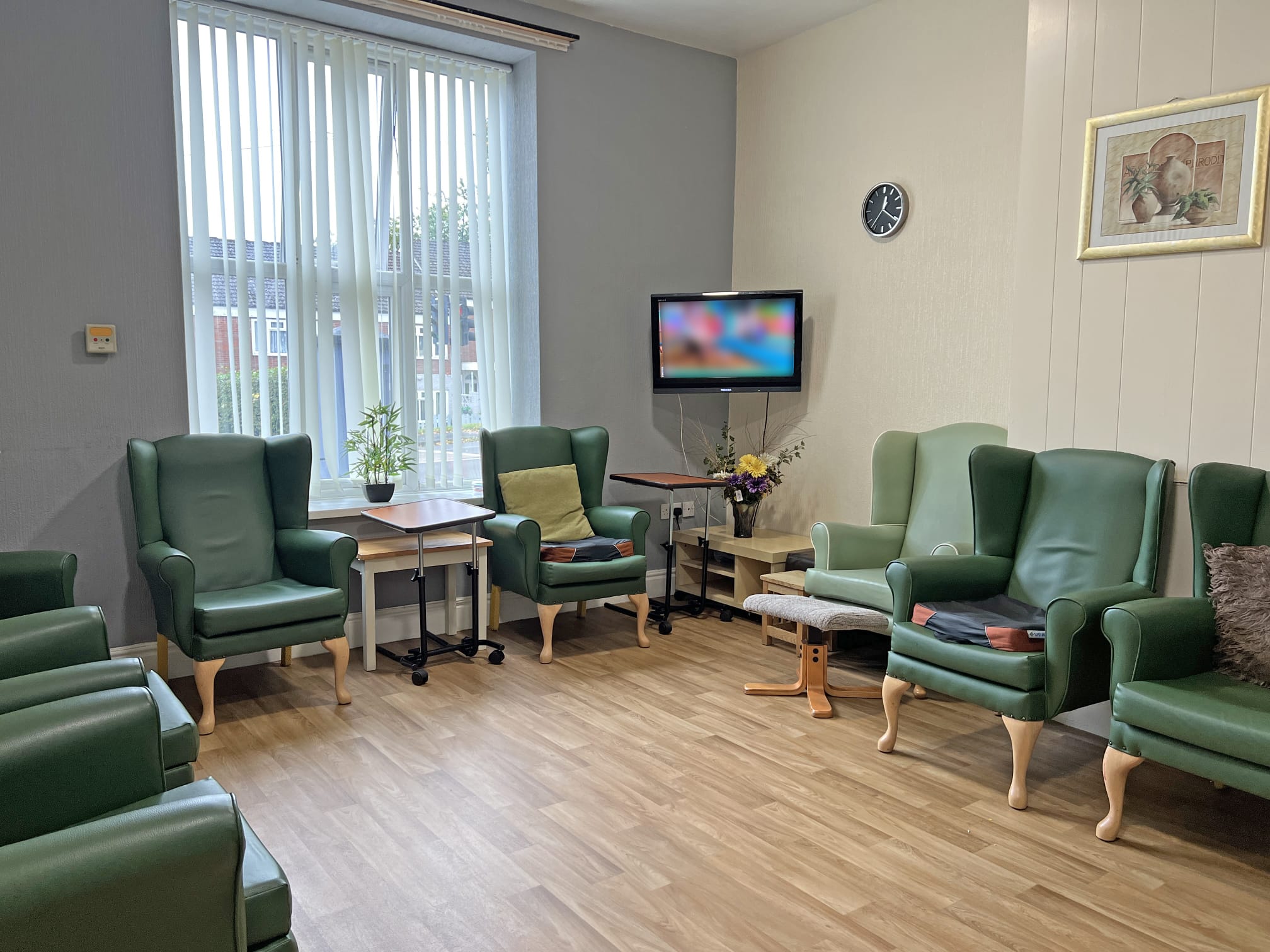 A bright, tidy room at Park Lane Care Home with green armchairs arranged in a circle, a window with vertical blinds, a TV on the wall, a clock, framed picture, small plant, and a vase of flowers on the table.