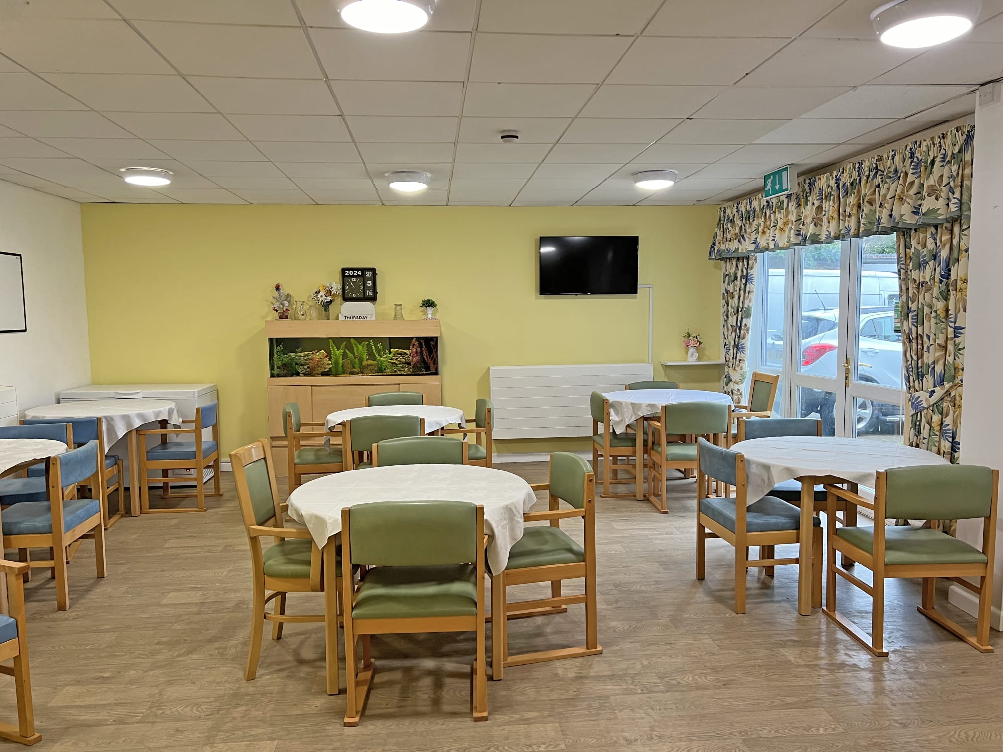 A bright, tidy dining room at Park Lane Care Home features round tables with white cloths, green-cushioned wooden chairs, a TV, fish tank, and floral curtains, with large windows letting in plenty of natural light.