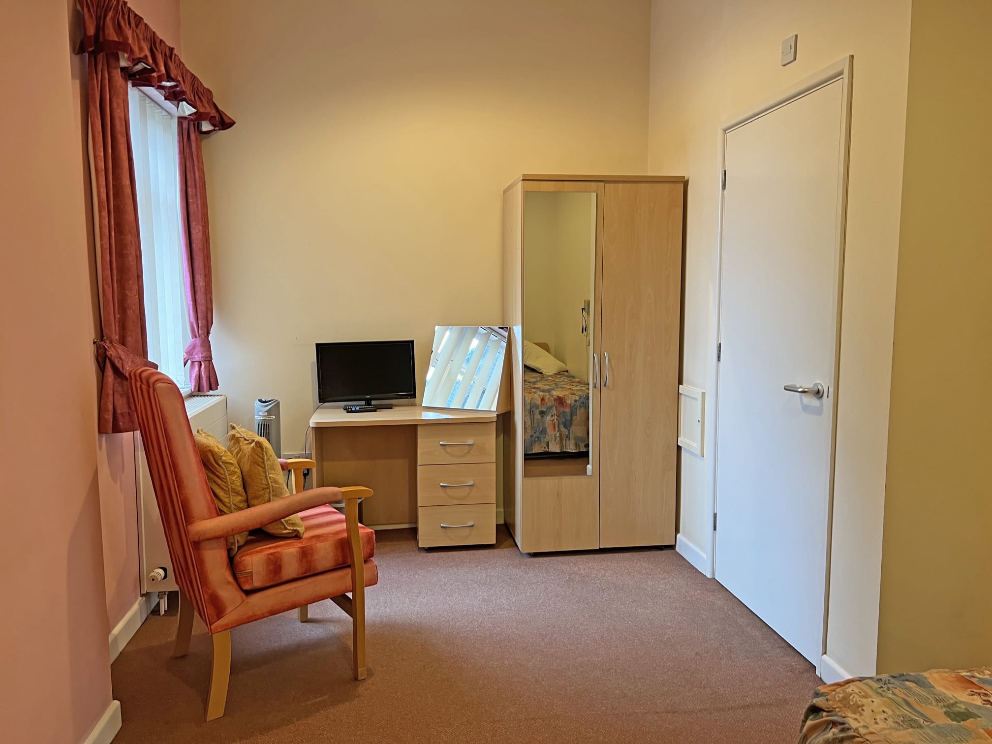 A tidy room at Park Lane Care Home features an orange-cushioned armchair, a window with red curtains, a desk with a small TV and mirror, a wardrobe with a mirrored door, and a closed white door on the right.