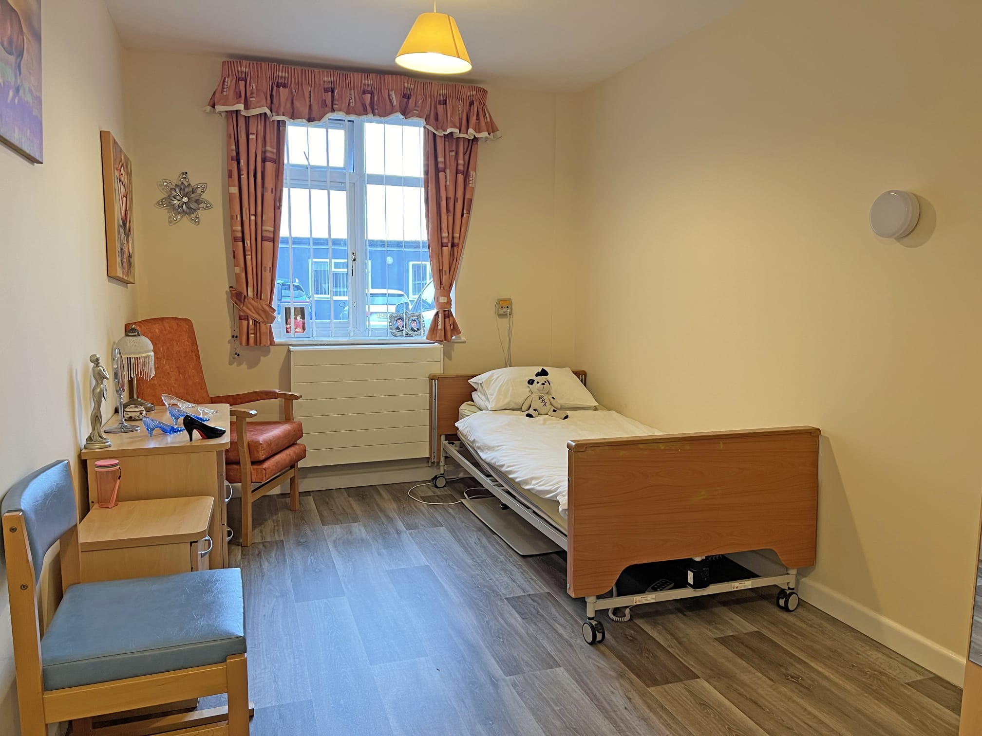 A tidy, single-occupancy room at Park Lane Care Home features a hospital bed, wooden furniture, an orange chair, and a window with patterned curtains letting in daylight. The floor is laminate, and the walls are pale yellow.