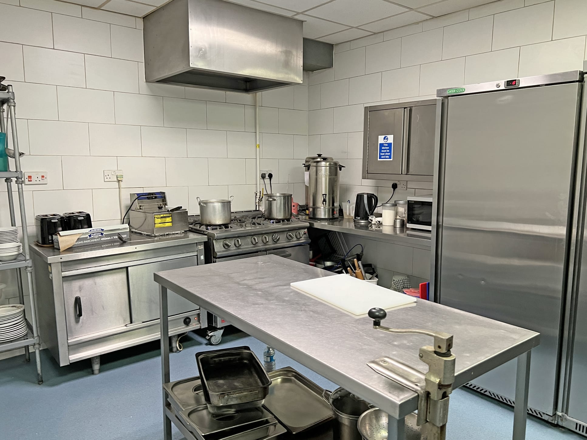 A commercial kitchen at Park Lane Care Home features a stainless steel table, cooking pots on a cooker, an industrial oven, a large fridge, and various kitchen equipment against white tiled walls.