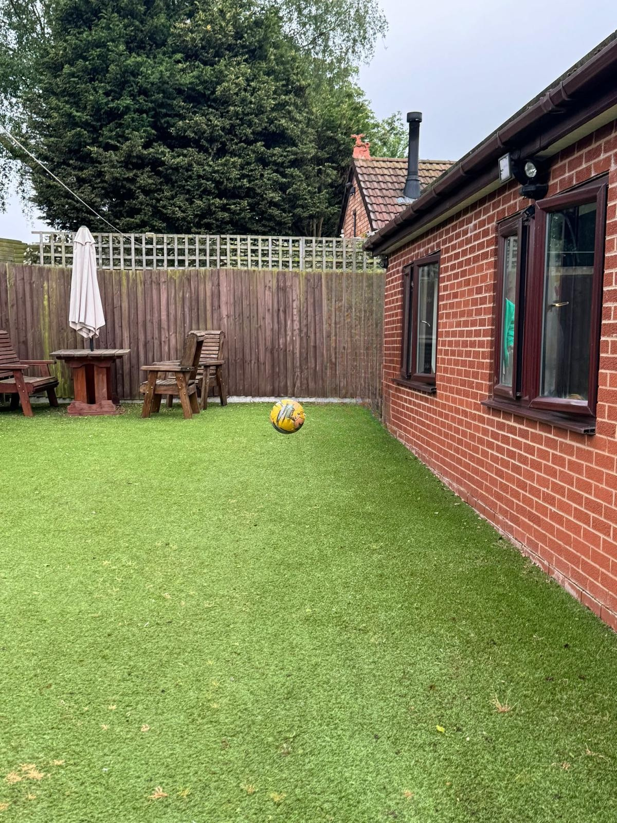 A yellow and blue football appears to float in mid-air on artificial grass near a residential home, with wooden garden furniture and a fence in the background.