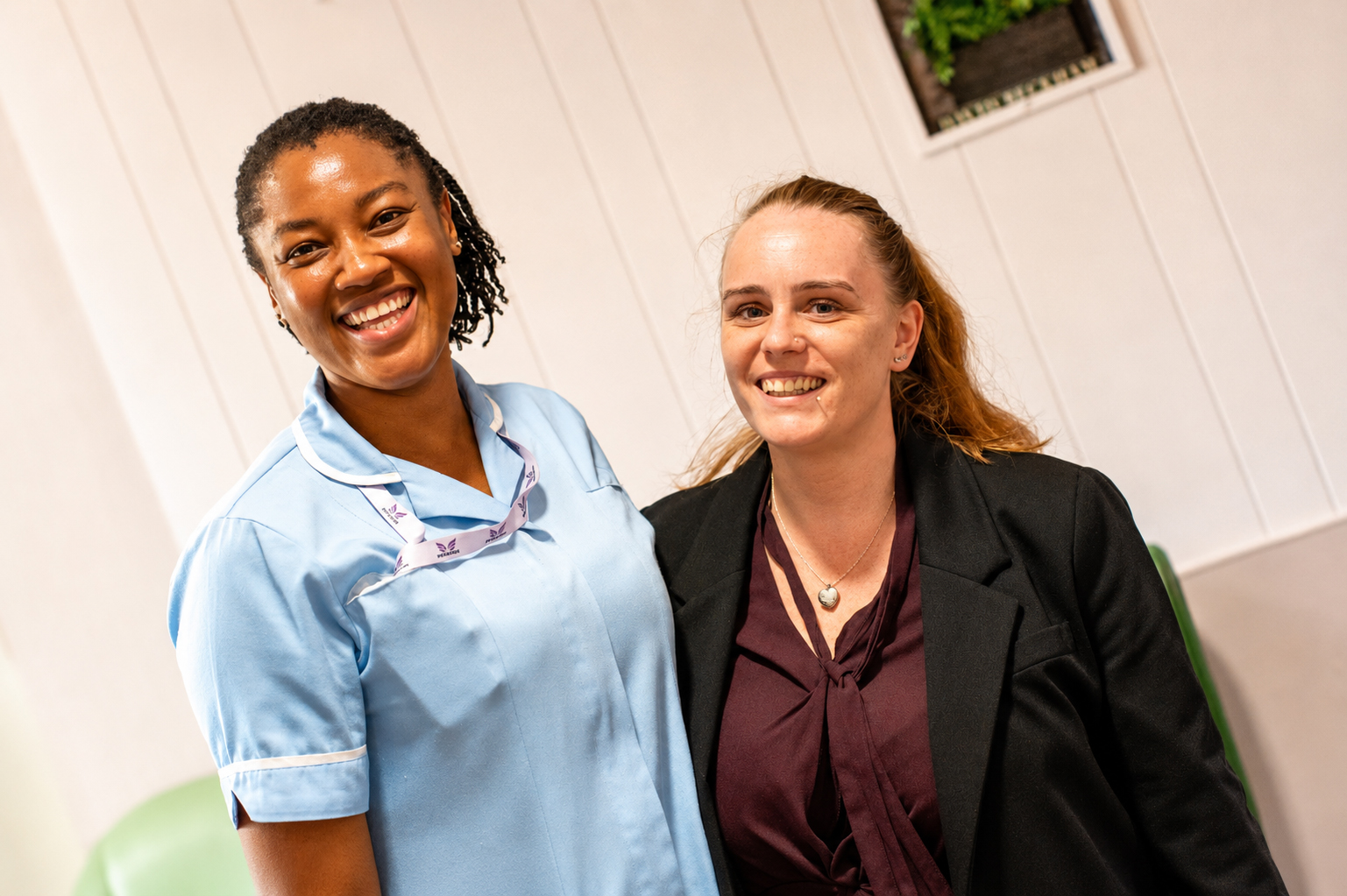Two women stand side by side smiling. One is wearing a light blue nurse uniform, and the other is in a dark blazer and maroon blouse. They are indoors, with a white wall and a small plant in the background.