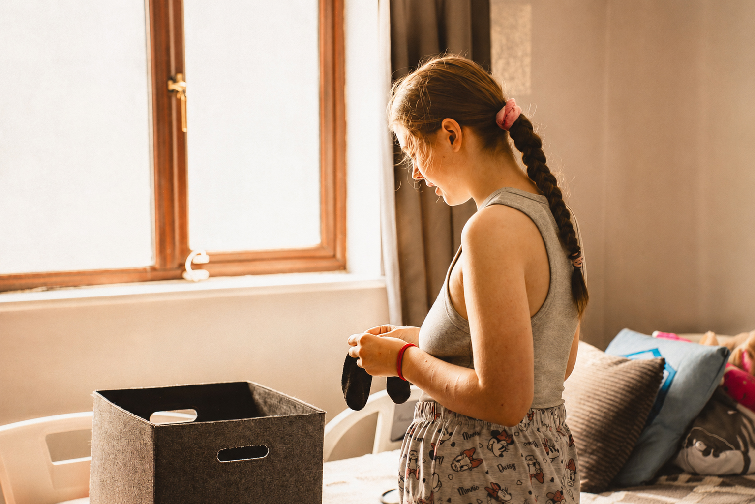 A woman with a plait stands by a window, folding clothes next to a fabric storage box on a bed in a sunlit room.
