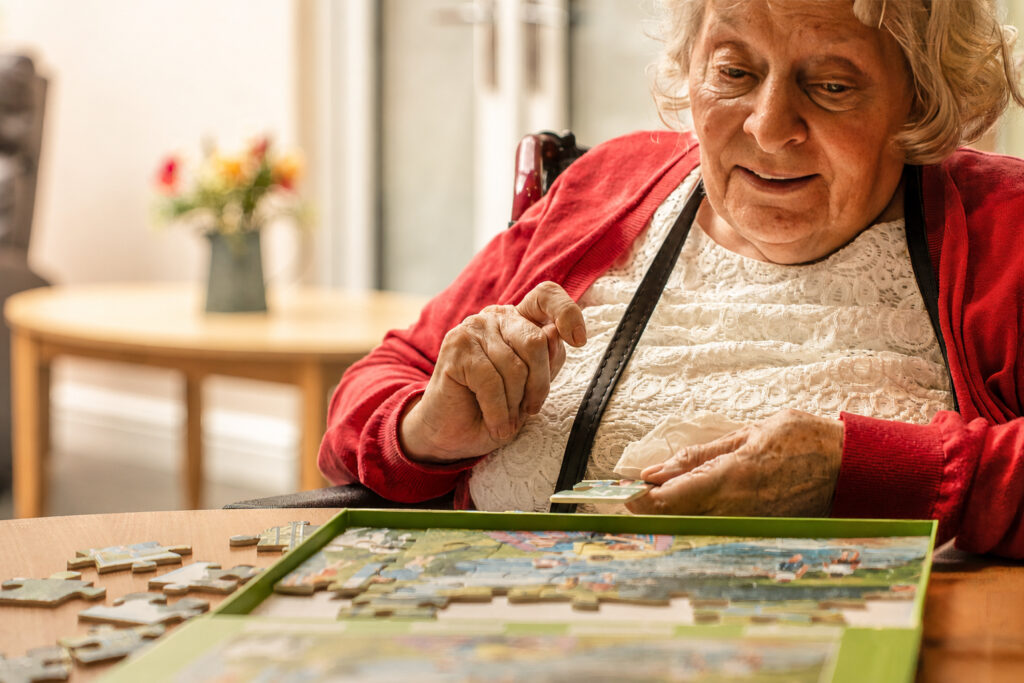 An elderly woman with grey hair and a red cardigan sits at a table, smiling whilst assembling a jigsaw puzzle. A vase of flowers is visible in the blurry background.