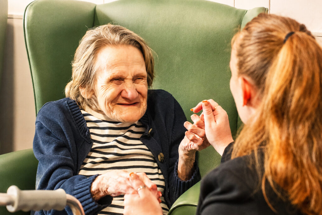An elderly woman in a striped shirt and blue cardigan sits in a green armchair, smiling and holding hands with a younger woman with reddish-brown hair, who faces her and smiles warmly.