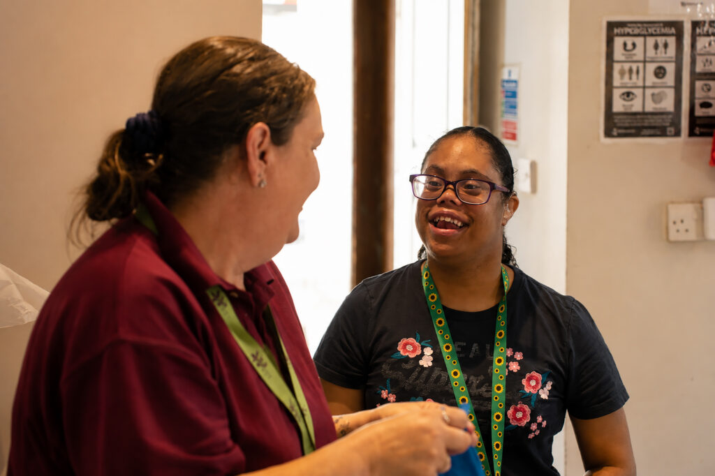 Two women at Harden Residential Home, wearing green lanyards, smile and talk indoors. One has a ponytail and burgundy shirt; the other wears glasses and a navy floral-embroidered shirt. Informational posters are visible on the wall behind them.