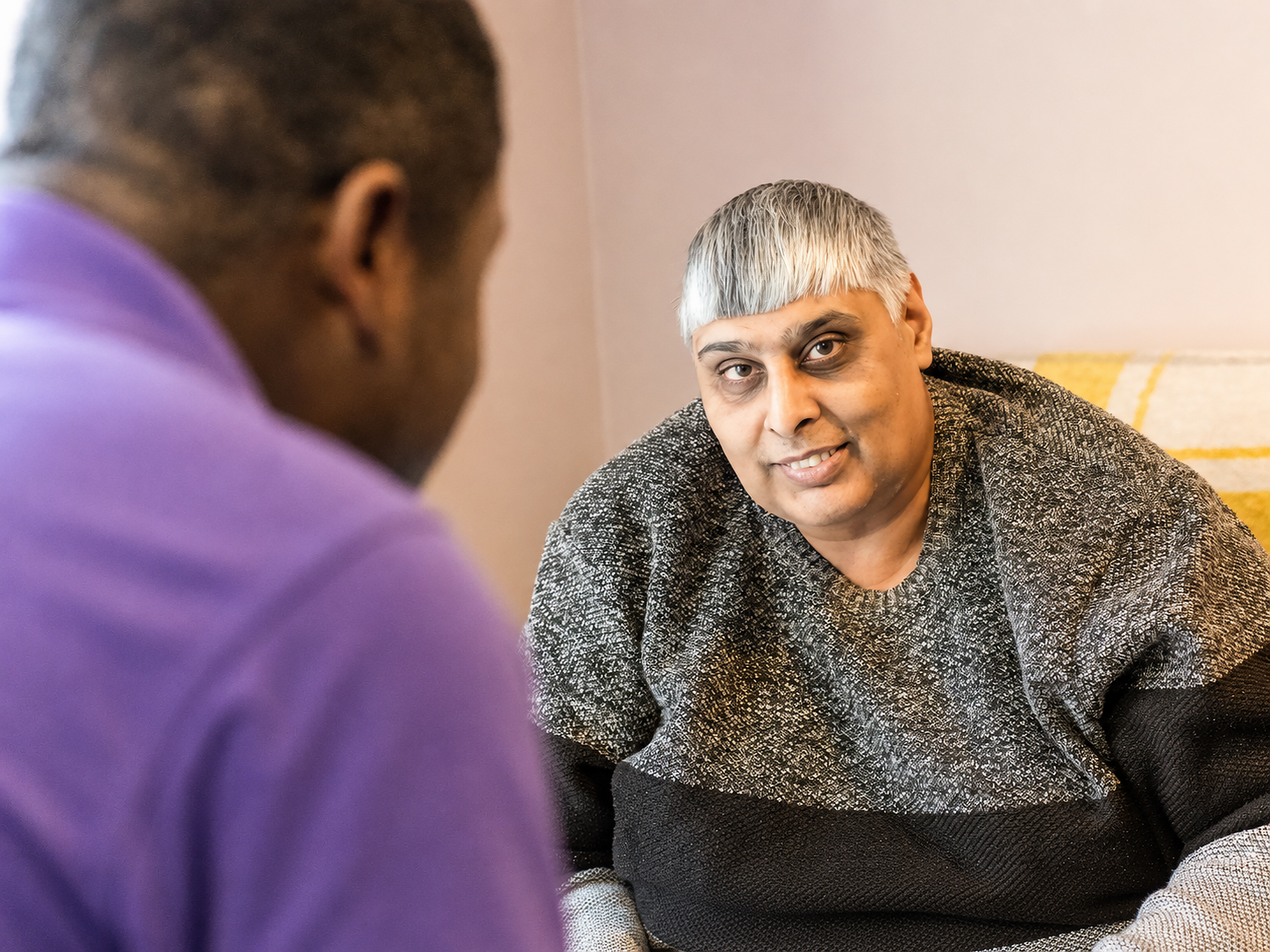 A person with short grey hair and a grey jumper sits on a sofa at Park Lane Care Home, looking at another person in a purple shirt who is turned away from the camera. They appear to be engaged in conversation.