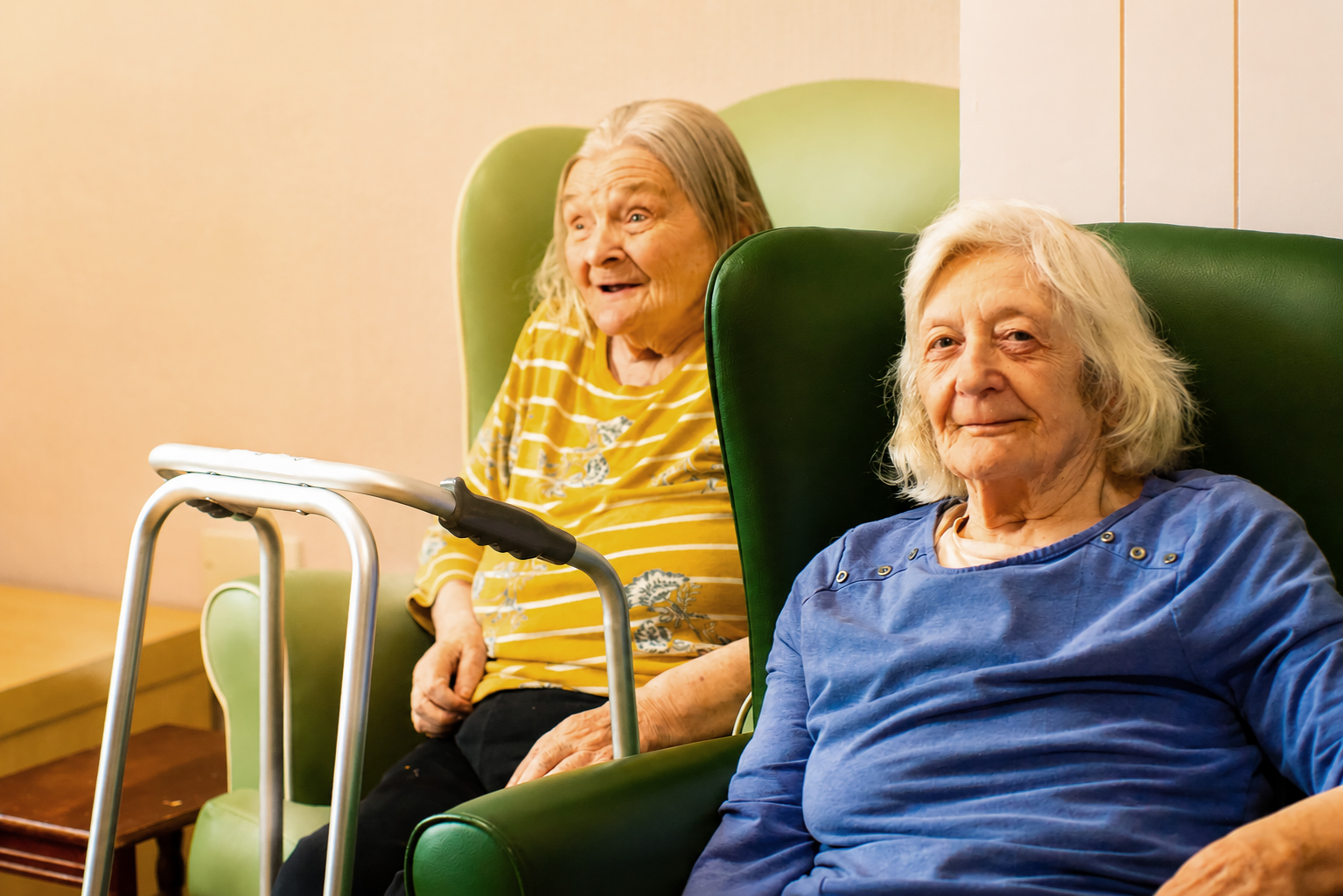 Two elderly women sit in green armchairs at Park Lane Care Home—one in a yellow striped shirt smiling with a walking frame nearby, the other in a blue shirt looking at the camera with a gentle expression.