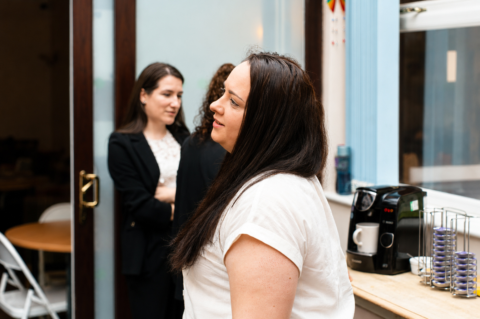 A woman in a white shirt stands near a coffee machine in a kitchen; two other women are talking in the background by a glass door.