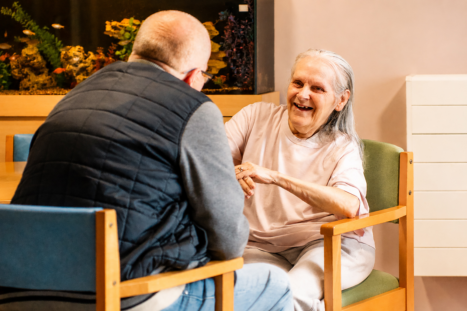 An elderly woman with long grey hair smiles and gestures while talking to a man in a black waistcoat, sitting opposite her in the brightly lit lounge of Park Lane Care Home, with an aquarium in the background.