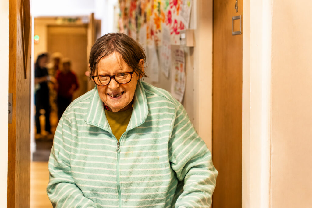 An older woman with glasses and short hair, wearing a green striped zip-up jumper, smiles whilst walking down a corridor at Park Lane Care Home decorated with colourful drawings.