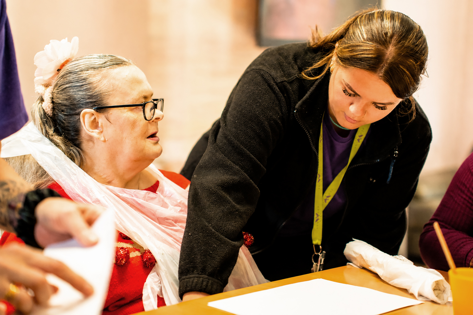 A carer at Park Lane Care Home assists an elderly woman wearing glasses and a flower in her hair, both looking at papers on a table in a well-lit room. The carer leans over to help with the activity.