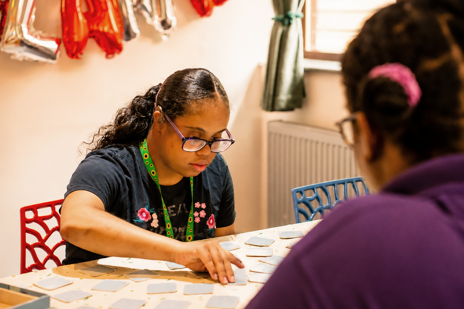 Two young women sit at a table playing a matching card game at Park Lane Care Home. One, wearing glasses and a dark shirt, is focused on choosing a card. The brightly lit room features colourful decorations and a window in the background.