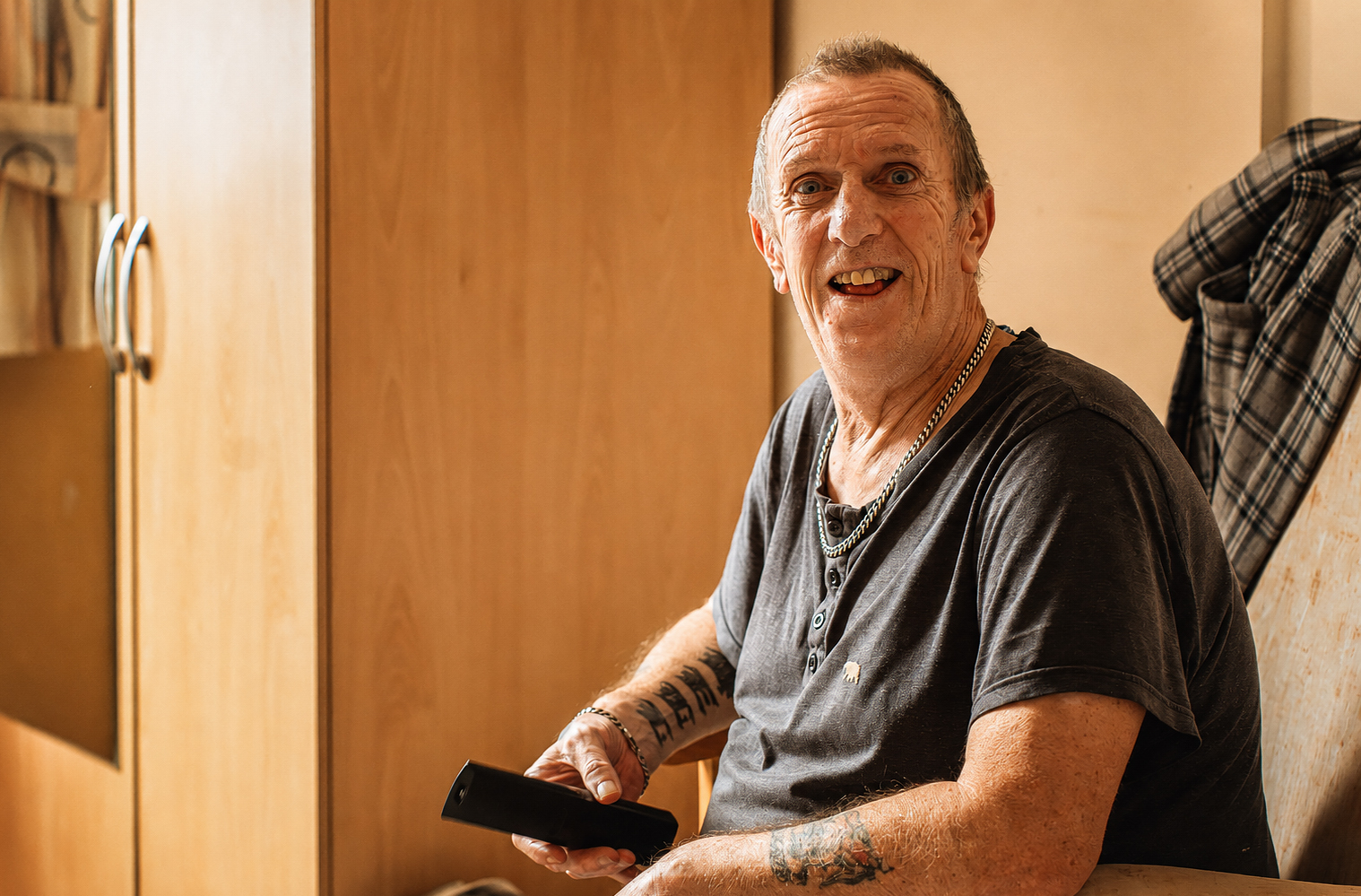 A smiling older man with tattoos sits in a chair at Park Lane Care Home, holding a TV remote. He wears a dark T-shirt and chain necklaces. A checked shirt hangs on the chair, with wooden cupboards in the background.