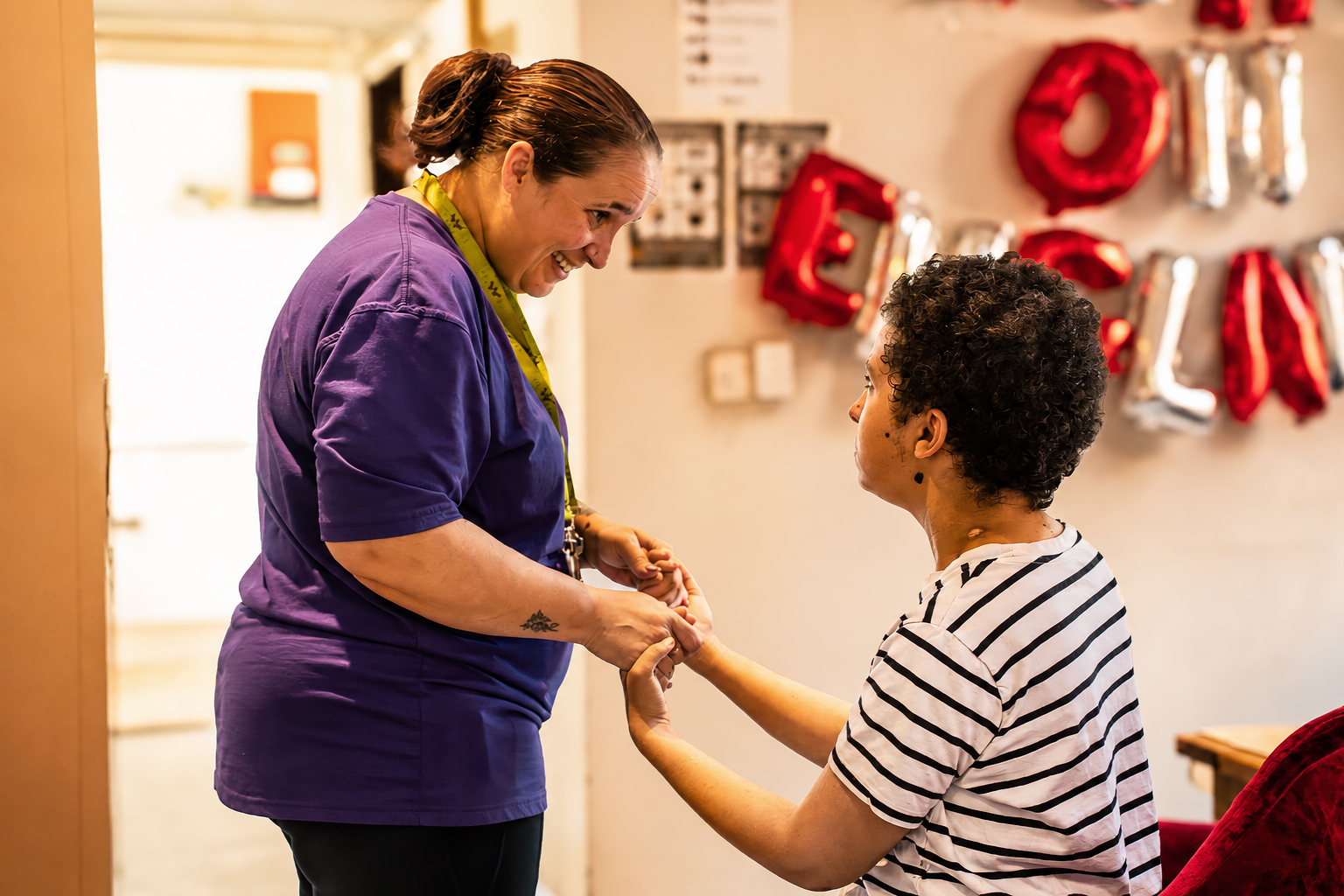A woman in a purple shirt smiles whilst holding hands with a seated person in a striped shirt at Park Lane Care Home. They appear to be engaging in a supportive or comforting interaction indoors, with red and silver balloons in the background.