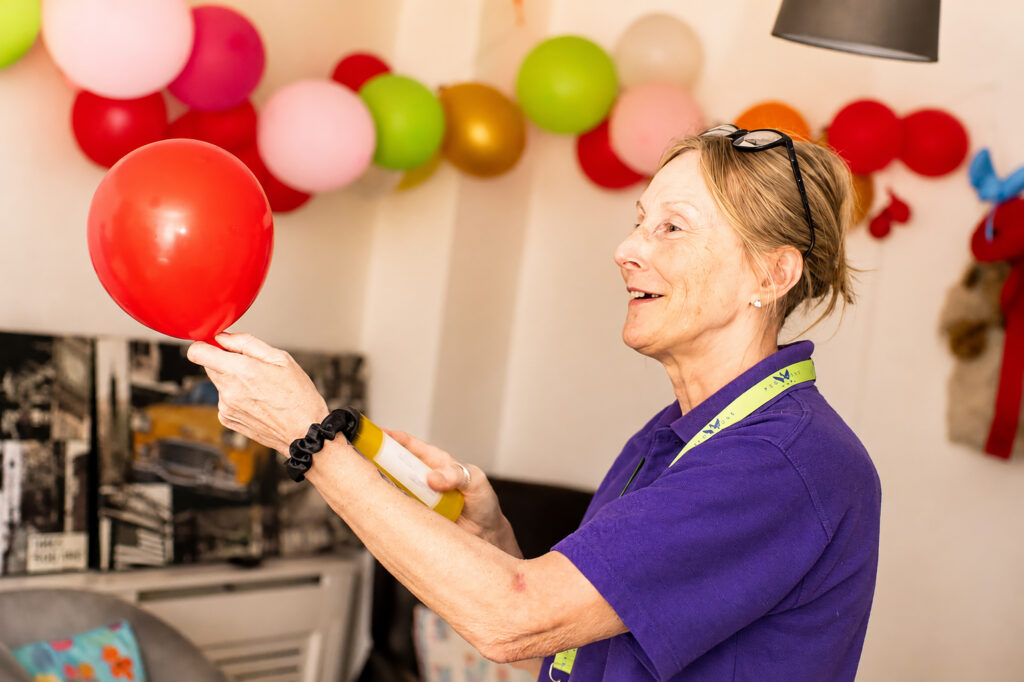 A woman in a purple shirt holds a red balloon, smiling. Colourful balloons and decorations hang on the wall behind her at Park Lane Care Home, creating a festive atmosphere indoors.