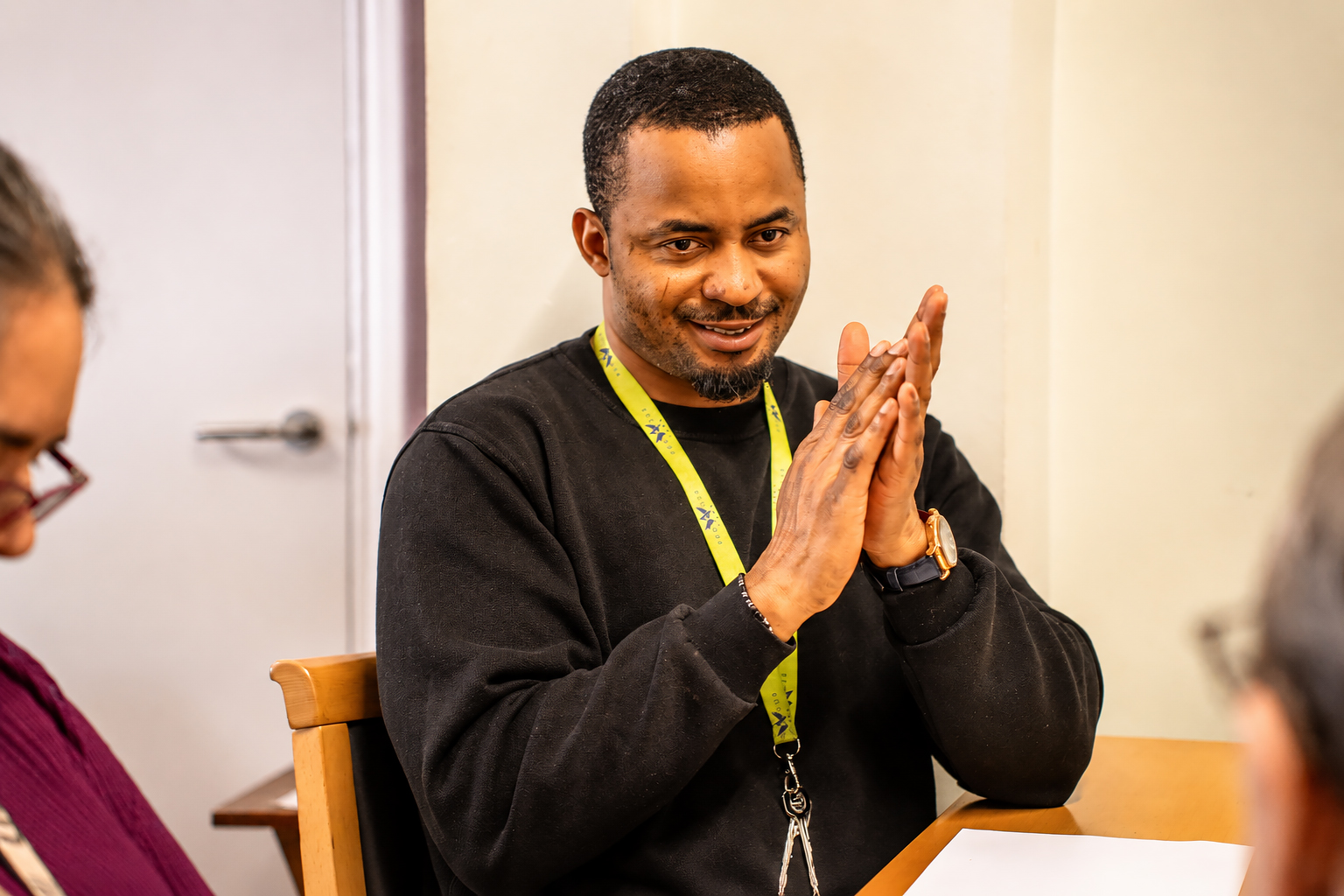 A man wearing a black jumper and yellow lanyard sits at a table, smiling and clasping his hands as he engages in conversation with others in the brightly lit Park Lane Care Home.