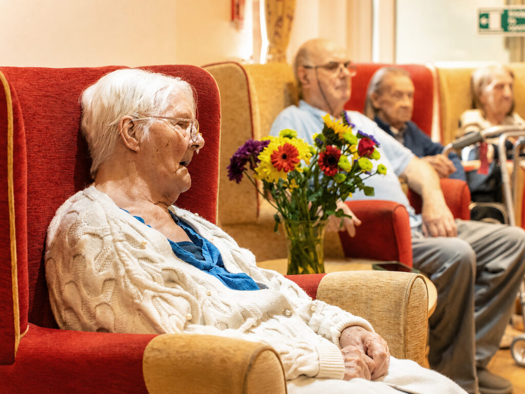 Elderly people sit in comfortable chairs at Park Lane Care Home. A woman wearing glasses and a white jumper is in the foreground, beside a vase of colourful flowers. Others enjoy the relaxed atmosphere in the background.