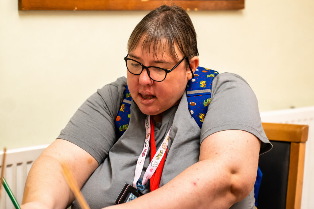 A person with short hair and glasses wears a grey shirt and a blue rucksack with cartoon characters. At Park Lane Care Home, they sit at a table, focussed on an activity, with a lanyard and name badge round their neck.