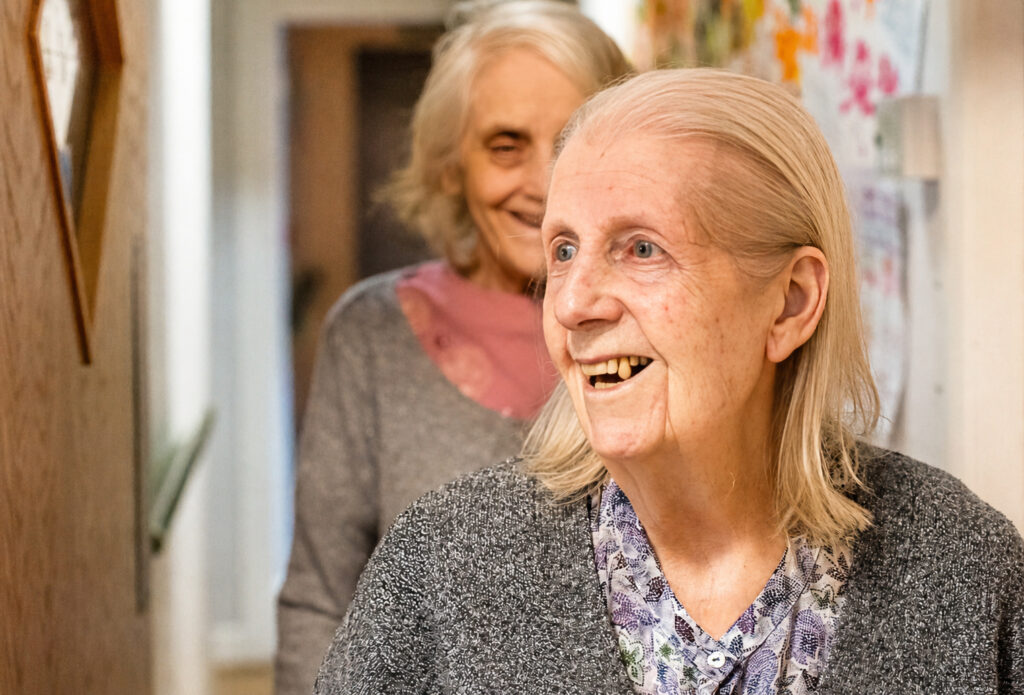 Two elderly women with fair hair and grey clothing smile warmly in a brightly lit corridor. The woman in front appears joyful, while the other stands slightly behind her, also smiling. Colourful drawings decorate the wall.