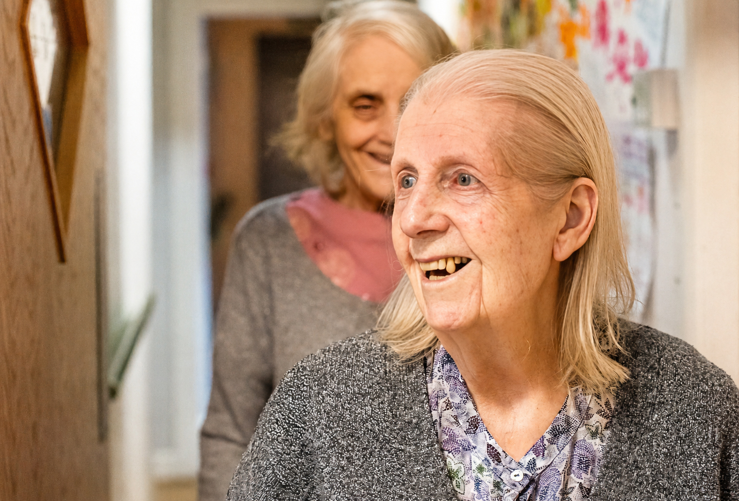Two elderly women with fair hair and grey clothing smile warmly in a brightly lit corridor. The woman in front appears joyful, while the other stands slightly behind her, also smiling. Colourful drawings decorate the wall.