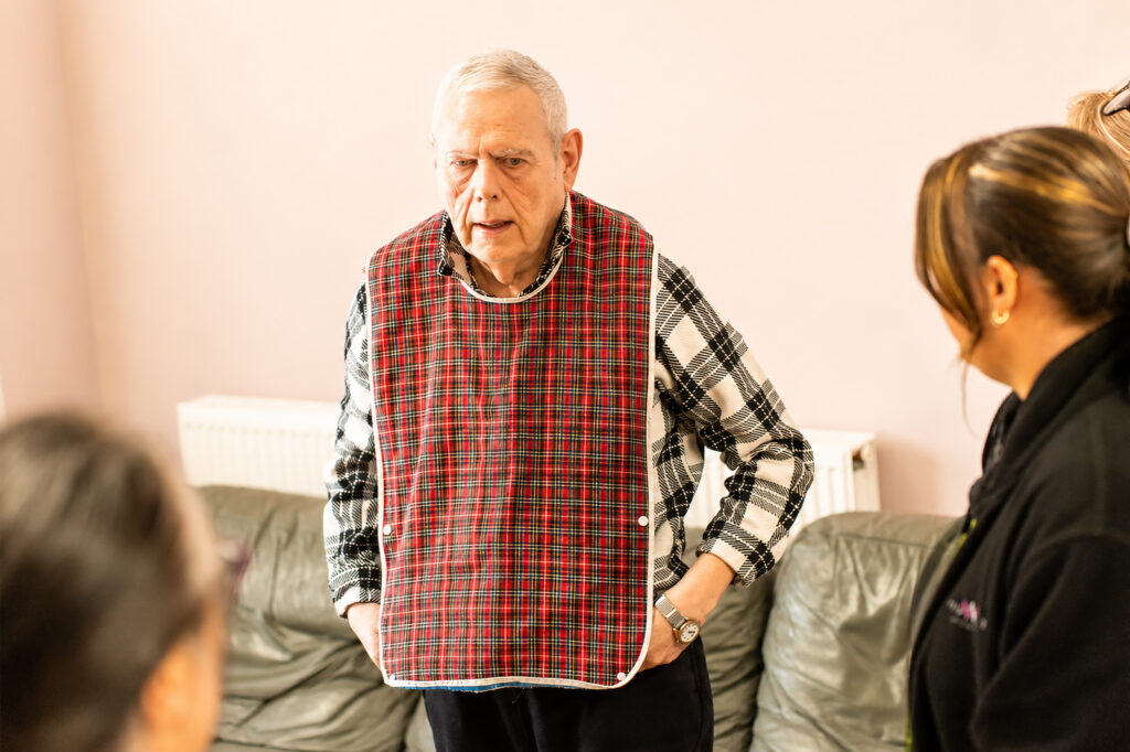 An older man wearing a checked adult bib stands in a lounge at Park Lane Care Home, facing three people who are partially visible in the foreground. He appears to be engaged in conversation with them.