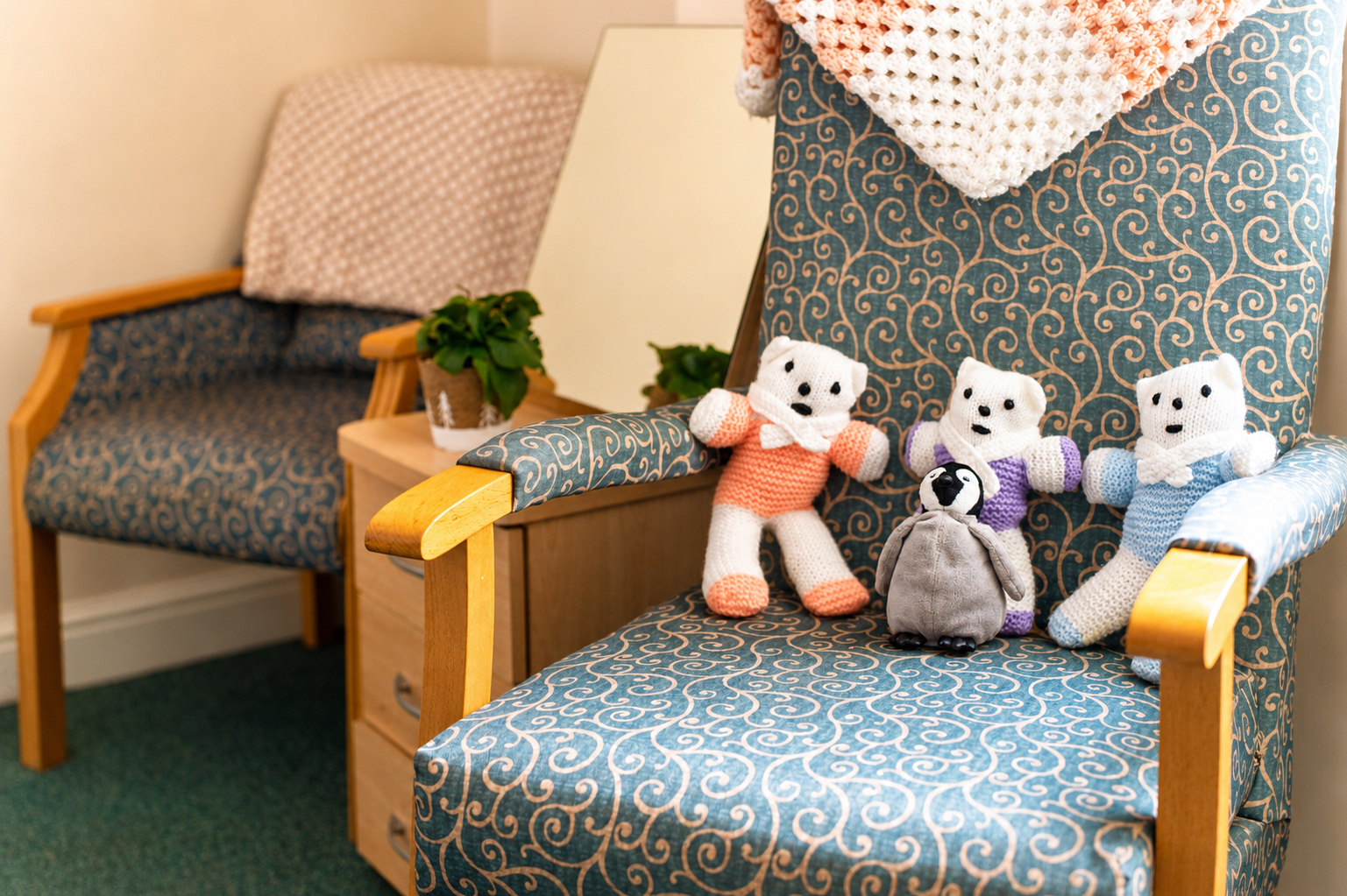 A patterned armchair with three knitted bear dolls and a small penguin soft toy on the seat. A crocheted blanket is draped over the back, and a second armchair and small plant are visible in the background.