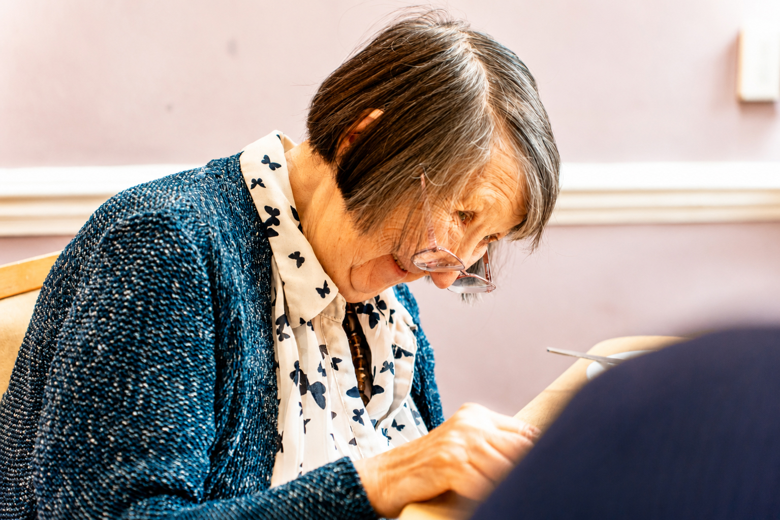 An elderly woman with short grey hair, wearing glasses, a blue cardigan, and a white blouse with butterfly prints, is sitting and concentrating on writing or reading at a table.