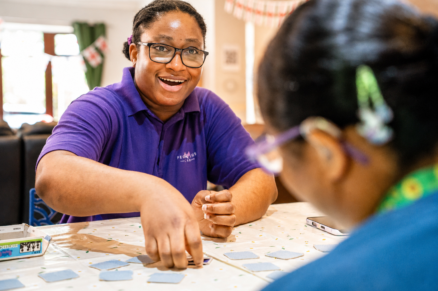 A smiling person in a purple polo shirt plays a card game at a table with another person, creating a cheerful and engaging atmosphere indoors.